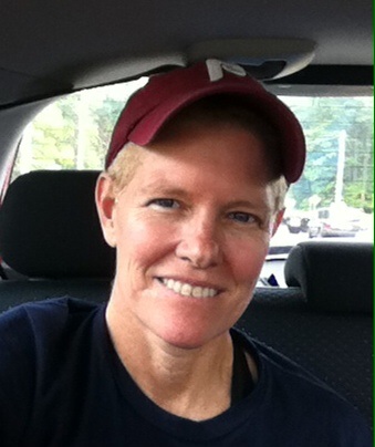 Headshot of person with short blonde hair, dark tshirt and red cap sitting in a car.