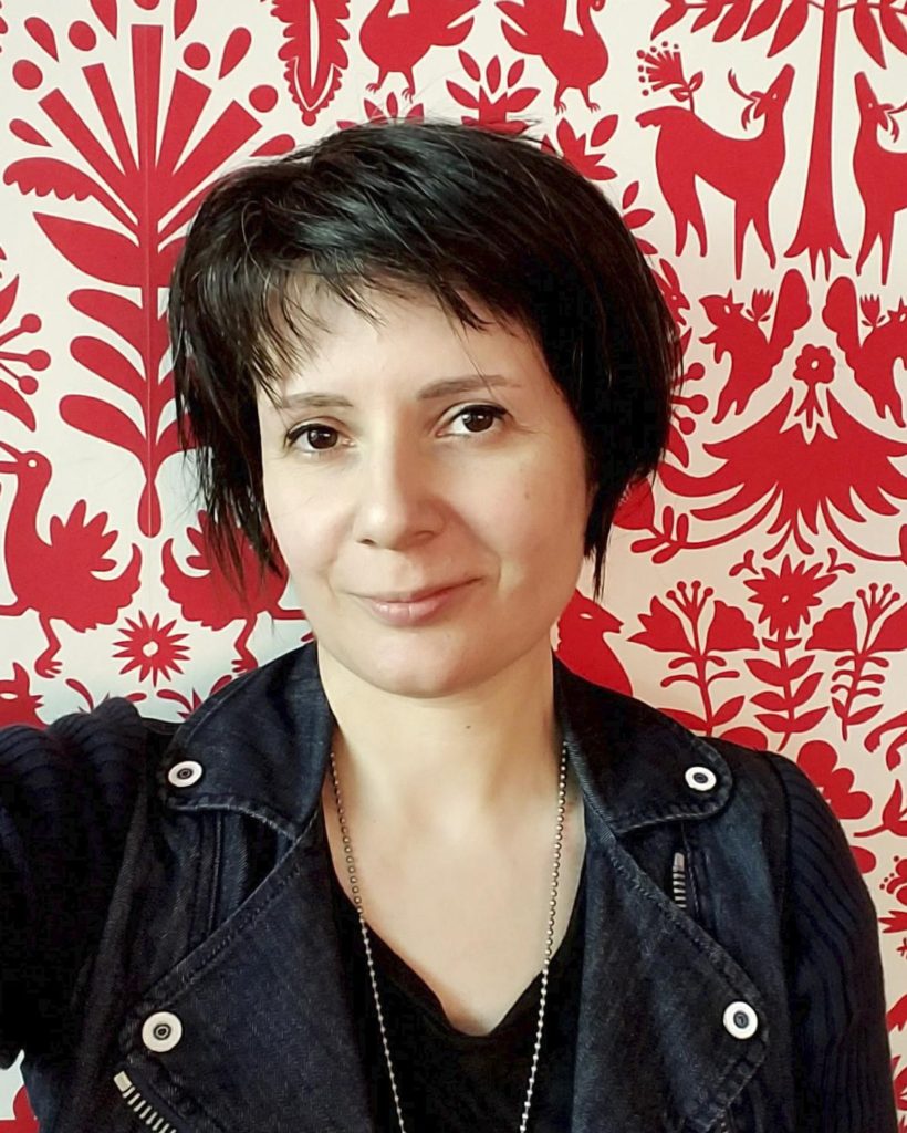Short haired person wearing a denim jacket looking straight into camera with red and white background.