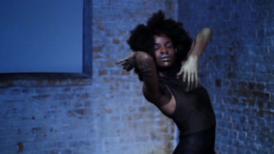 A black person with an afro and wearing a tight black outfit is dancing, appearing absorbed in the moment. In the background is a brick wall. All is bathed in blue light.