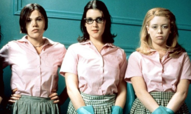 Three people stand in a row looking bored and at the camera: a white person with short dark hair, a white person with long dark hair and glasses, and a white person with long blonde hair. They are all wearing an apparent uniform of pink blouses green pinnies, and bright blue wasing up gloves. In the background is a bright turquoise wall.