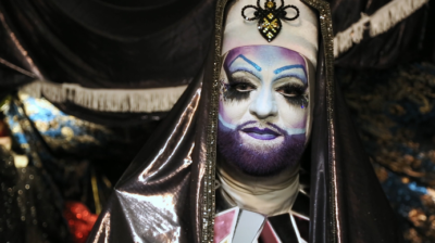 A white person wears an elaborate habit made of shiny material and a jeweled bee design on the forehead. They wear white make-up covering their face with a purple beard and lips, and the blue and pink of the trans flag elsewhere on their face plus big black eye make-up.