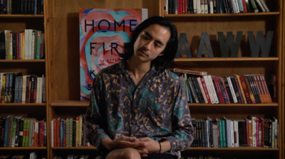 An Asian-American man sits wtih legs crossed, his head titled thoughtfully to one side, looking down. He has long black hair and wears a patterned shirt. In the background are bookshelves.