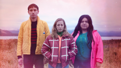 A white man with short dark hair and a yellow jacket, white woman with long blonde hair and a red and white striped jacket, and Latina woman with long dark hair and luminous pink jacket stand in a field looking directly at the camera.