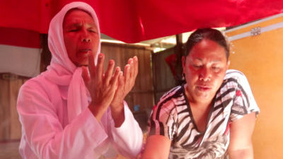 Two Bugis women from southern Indonesia sit next to each other in a wooden building with red awning. One has a white shirt and headscarf and appears to be in prayer. The other woman wears a black and white tshirt with a tiger on it and is looking down.
