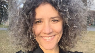 Close up of a white woman with curly grey hair, smiling. Behind her grass is visible.