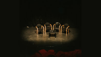 Four black and wooden chairs and a small black table sit ominously in a spotlight surrounded by black.