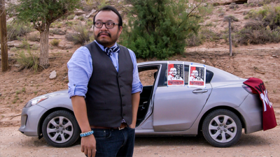 A brown man in light purple shirt, waistcoat, and tie with short dark hair, beard, and glasses stand with his hand in his pocket, looking up to the side. Behind him is a silver car with election posters on it, desert, and trees.