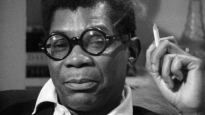 Black and white close up of a black man, Jason Holliday, gazing directly at the camera with a sly perhaps annoyed look. He has short black hair and glasses, and holds a cigarette in his hand.