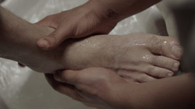 Close-up of one set of hands washing the foot of another person with water in a basin below.