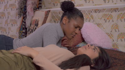A black person with hair up in a bun and blue jumper lies on a bed leaning over a brown person with long dark hair and a pink jumper. There is a pink cushion and flowery wallpaper behind them.