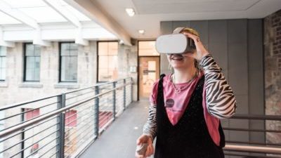 A white person with blond hair, wearing a pink SQIFF tshirt with zebra-stripped long-sleeve tshirt under it and black dungerees has a virtual reality headset on. They are upstairs at the CCA.