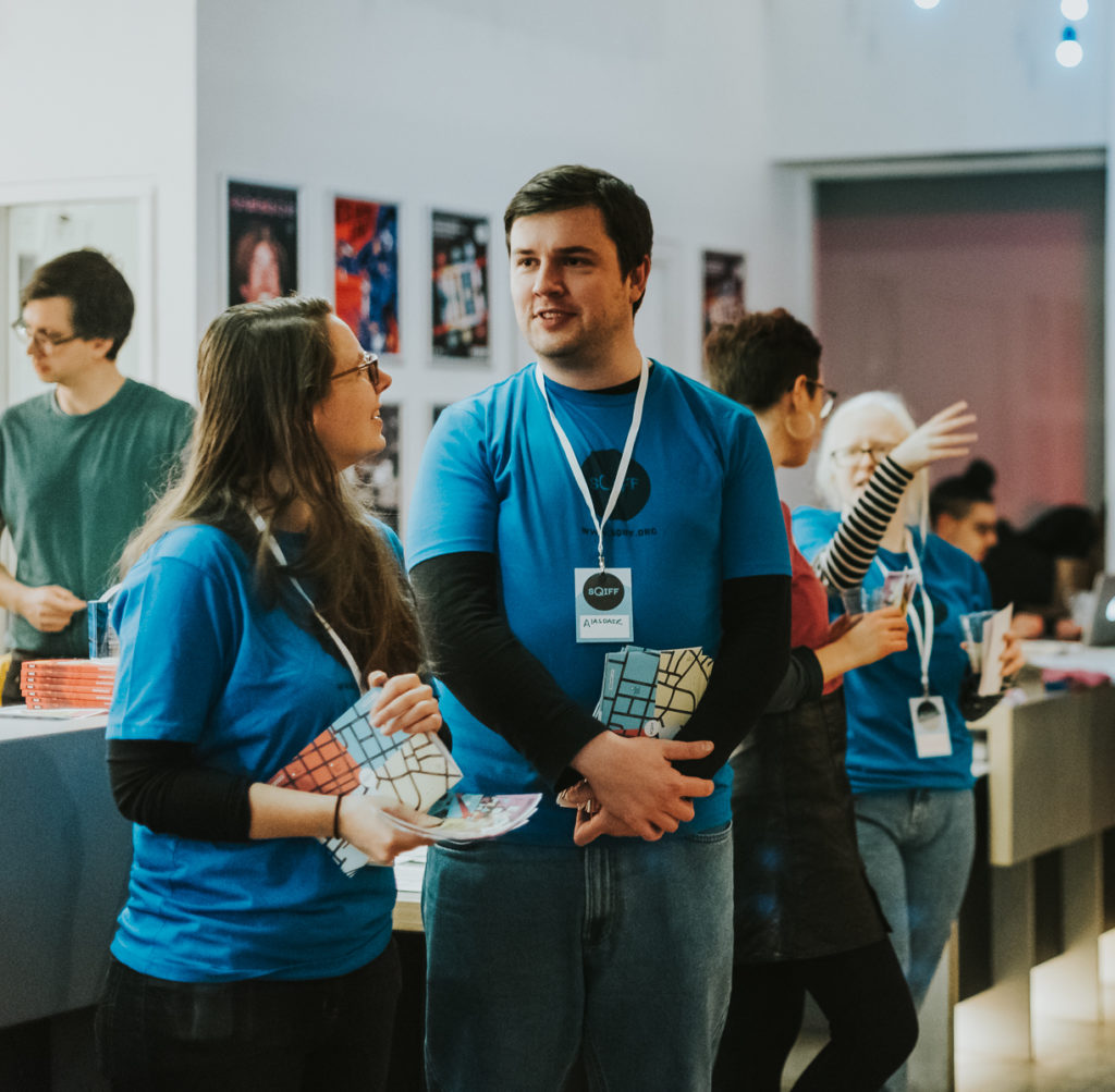 Two white people with blue SQIFF tshirts holding a handful of SQIFF brochures each stand in the foreground of the CCA foyer with people milling behind them.