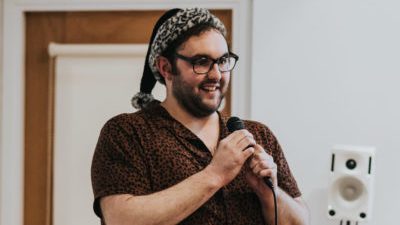 A white person with short dark hair, a beard, glasses, and wearing a snow leopard santa hat grins, speaking into a microphone.