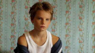 A white child with short, messy blond hair, wearing a white vest and blue tracksuit top falling off their shoulders, stares sullenly ahead. A wall with light green and pink flowered wallpaper is behind them.