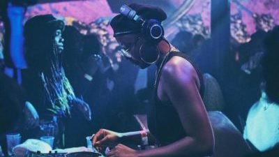 A black person with a black vest, hat, and big headphones is DJing with a crowd of people behind.