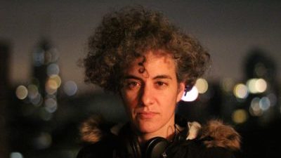 A white person with big curly mousy hair, headphones round their neck, and a big jacket looks direct to camera. Behind thmem are blurred city lights.