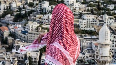 A figure with a pink headscarf covering their top half completely with their back to camera looks out across a bright city filled with tall white buildings.