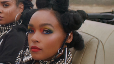 A black woman - Janelle Monae - is in close up looking just beyond the camera. She has black hair in pigtails and wears multi-coloured eye make-up, bright red lipstic, and a pearl collar. Just behind her another black woman can be seen. They are both sitting in an open top car.