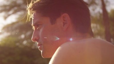 Close up of the head and naked shoulders of a white man with short dark hair. He is looking whistfully to the side and his hair is caught by sunlight also coming through the tree tops behind him.