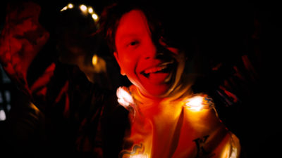 A white person bathed in red light and shadow looms out of the dark, grinning with light playing about them.