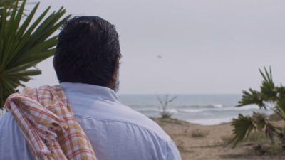 A brown man with his back to camera, short dark hair, a white shirt, and red and white checked clothing item over his shoulder, looks out to sea from a sandy beach with palm trees.