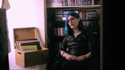 A white person with long blue hair and glasses, a black leather jacket, sitting down. There is a bookshelf with DVDs and CDs behind them and an open box full of records to their right.