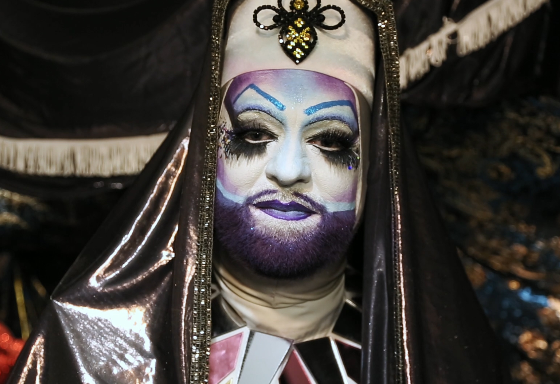 A white person wears an elaborate habit made of shiny material and a jeweled bee design on the forehead. They wear white make-up covering their face with a purple beard and lips, and the blue and pink of the trans flag elsewhere on their face plus big black eye make-up.