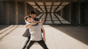 Two people stand legs apart in a dancing pose surrounded by concrete, behind them a reflection caused by sunlight and windows. One person with short black hair and a dark jacket and trousers has their arm around the neck of the other person, who has short dark hair, a grey tshirt, and dark trousers, in dance pose.