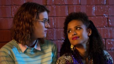 A still from the Black Mirror episode San Junipero: a white woman with long frizzy blonde hair, glasses, a shirt, and stripy jumper gazes at a black woman with long black hair, gold earrings, and a gold top. Behind is a brick wall.
