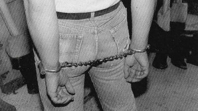 Black and white grainy archive image of a man with white tshirt and tight jeans with his hands handcuffed behind his back.