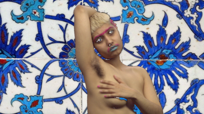 A brown person with short bleached blond hair, pink eyebrow make-up, and light blue lipstick poses, their upper half naked, one hand over their chest and one above their head. Behind them is a blue flowered pattern.