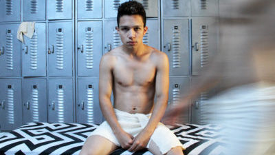 A white young person starring intensely ahead with short dark hair sits in a changing room wearing just a small white towel around his waist as another white person with a white towel is seen walking part, blurred.
