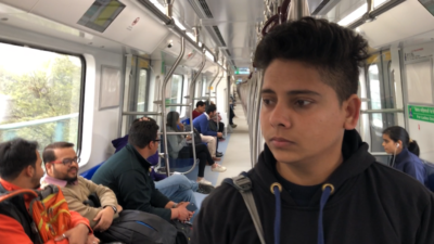 A brown person with short dark hair, a dark hoodie, and bag over their shoulder stands in a subway carriage looking down to the side with people sitting behind them.