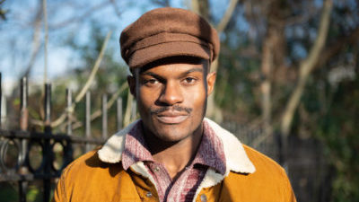 A black man looks directly at the camera. He has a small moustache and is wearing a brown flat cap, red and white shirt, and tan with white collar jacket. Behind him is a park railing and trees.