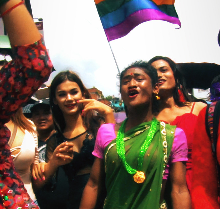 Still from short film Gai Jatra in which a Hindi person wearing a green sari is surrounded by other celebrating, one waving a rainbow flag.