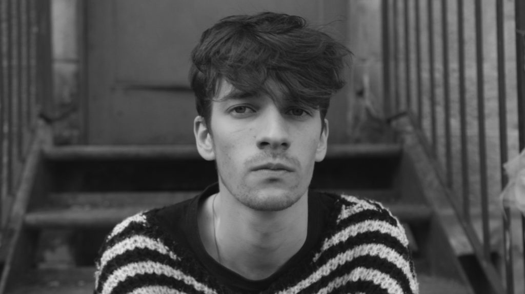 Sean Lìonadh, a white man with short dark hair wearing a black and white striped jumper, stares moodily at the camera.