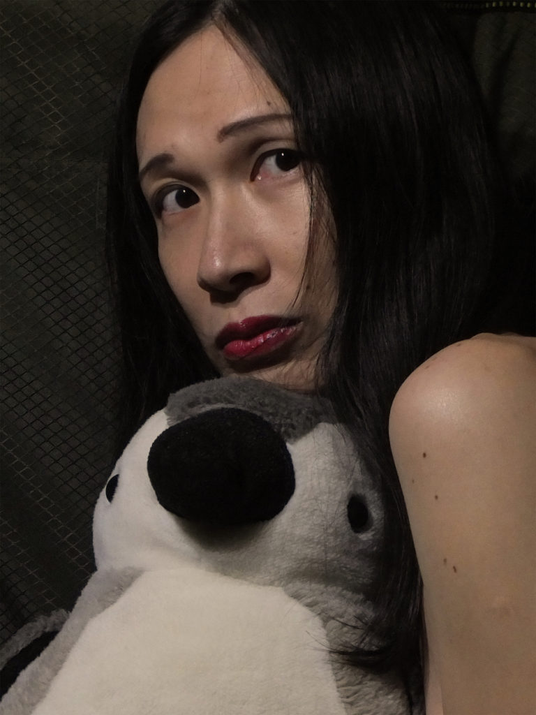 An East Asian woman, Beatrice Wong, stares at the camera with bare shoulder showing, a toy penguin, and surrounded by black.