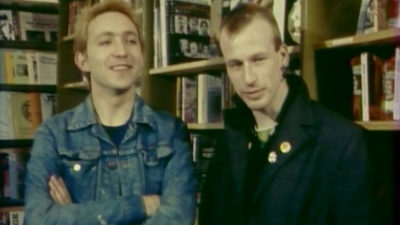 Grainy image of two men standing in front of book shelves. They both have short blonde hair. One wears a demin jacket and is crossing his arms and smiling. The other wears a black jacket with collar turned up and is looking to the side.