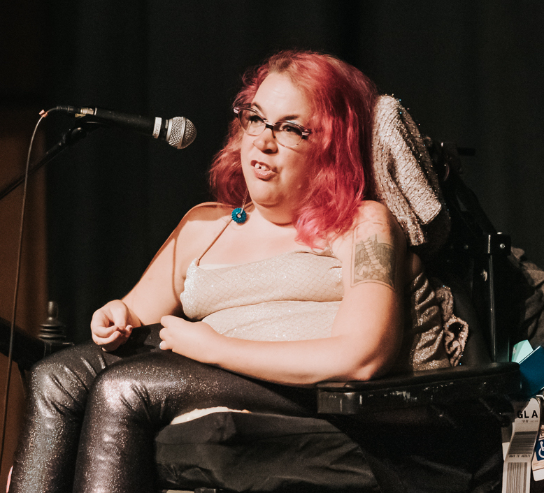 A woman with red hair, glasses, white vest, and leather trousers sits in her wheelchair, talking into a microphone.