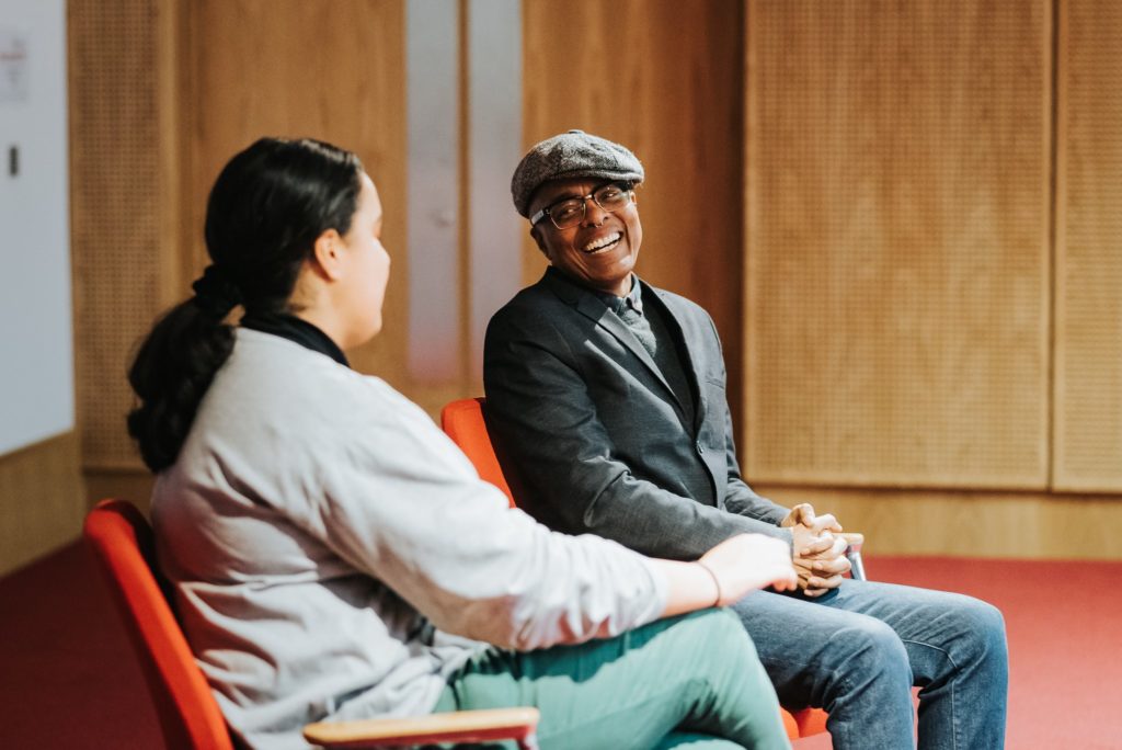 Two people sit in chairs at the front of a cinema space. One wears a flat cap and suit jacket and is grinning at the other, who has their back to us.