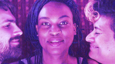 A Black person with braids and a slight grin is in close-up looking directly to camera. On either side looking at them and smiling are a White person with a dark beard and a brown person with a bleached forelock and nose ring. All are bathed in pink and purple light.