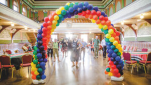 A ballroom filled with people being led in a dance with tables and chairs round the side and a rainbow arch made of balloons at the front framing them.