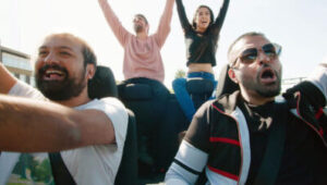 Four people are in a moving, open topped car with their hands in the air, smiling and shouting in a celebratory way.