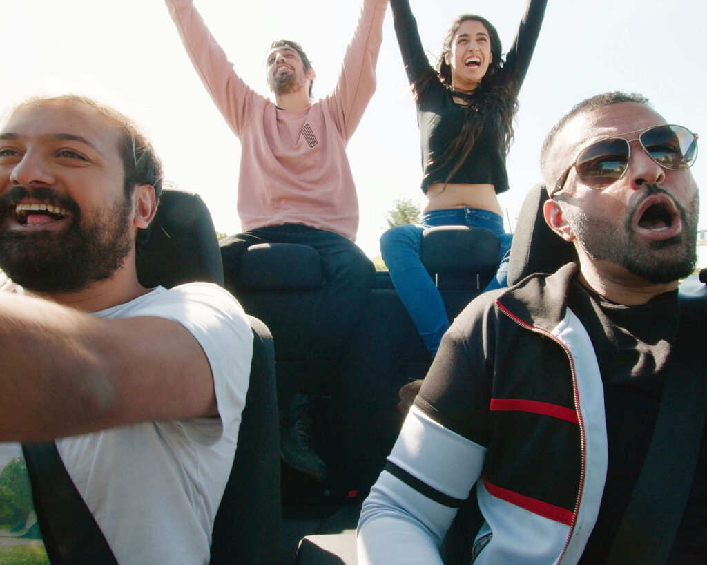 Four people are in a moving, open topped car with their hands in the air, smiling and shouting in a celebratory way.