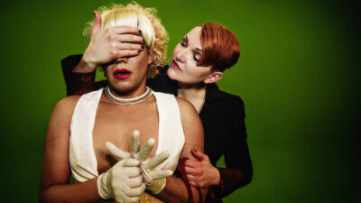 A white person with short red hair wearing a black suit jacket stands with her hand over the eyes of a second white person with blond hair in curls, red lipstick, a white bra-crop top, and rubber gloves.