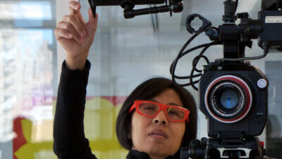 Lana Lin, a woman of colour with short black hair, bright red glasses, and a black top is standing next to a large camera with her hand up adjusting the monitor.