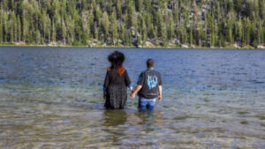Two people, one with long bright red hair and dramatic black attire and one with short hair dressed butch casual, stand hand in hand knee deep in a lake with a hill filled with trees beyond it.