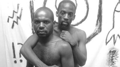 Two Black men are naked from the waist up in front of a white sheet with sketches of rocket and other items on it. One has an arm around the other's shoulder and they both look directly at the camera.