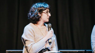 An Arab woman with curly black hair, glasses, and a woolly jumper sits mid-talking holding up a mic with a black wall behind her.