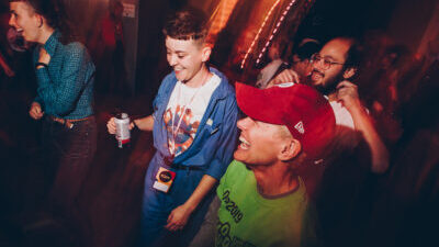 A group of queer people are grinning and having fun on a dark dancefloor with blurry red and pink lights.